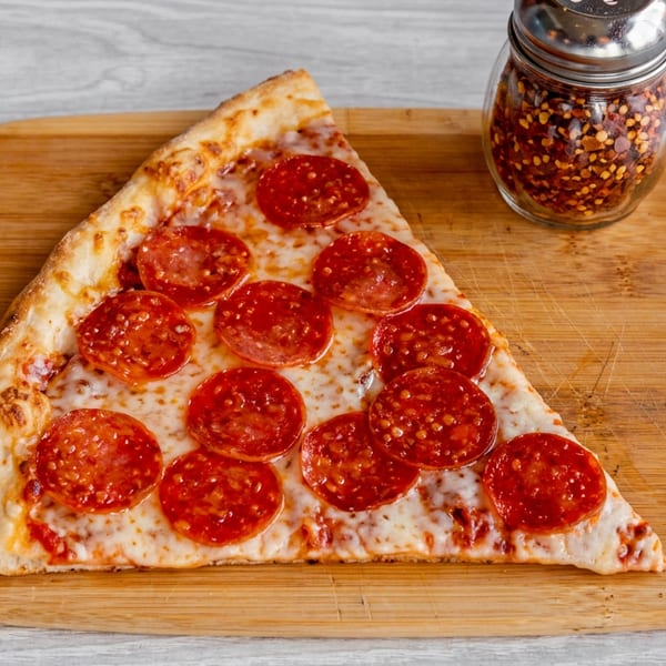 A slice of pepperoni pizza and a shaker of red pepper flakes on a wooden cutting board.