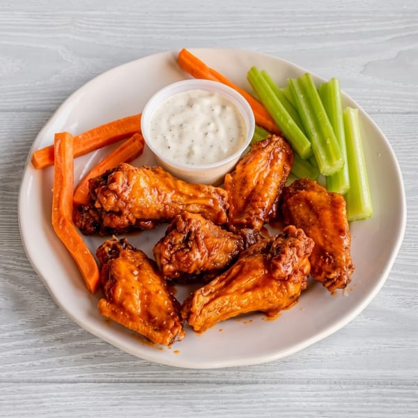A plate of buffalo wings with ranch dressing, carrot sticks, and celery on a white wooden table.